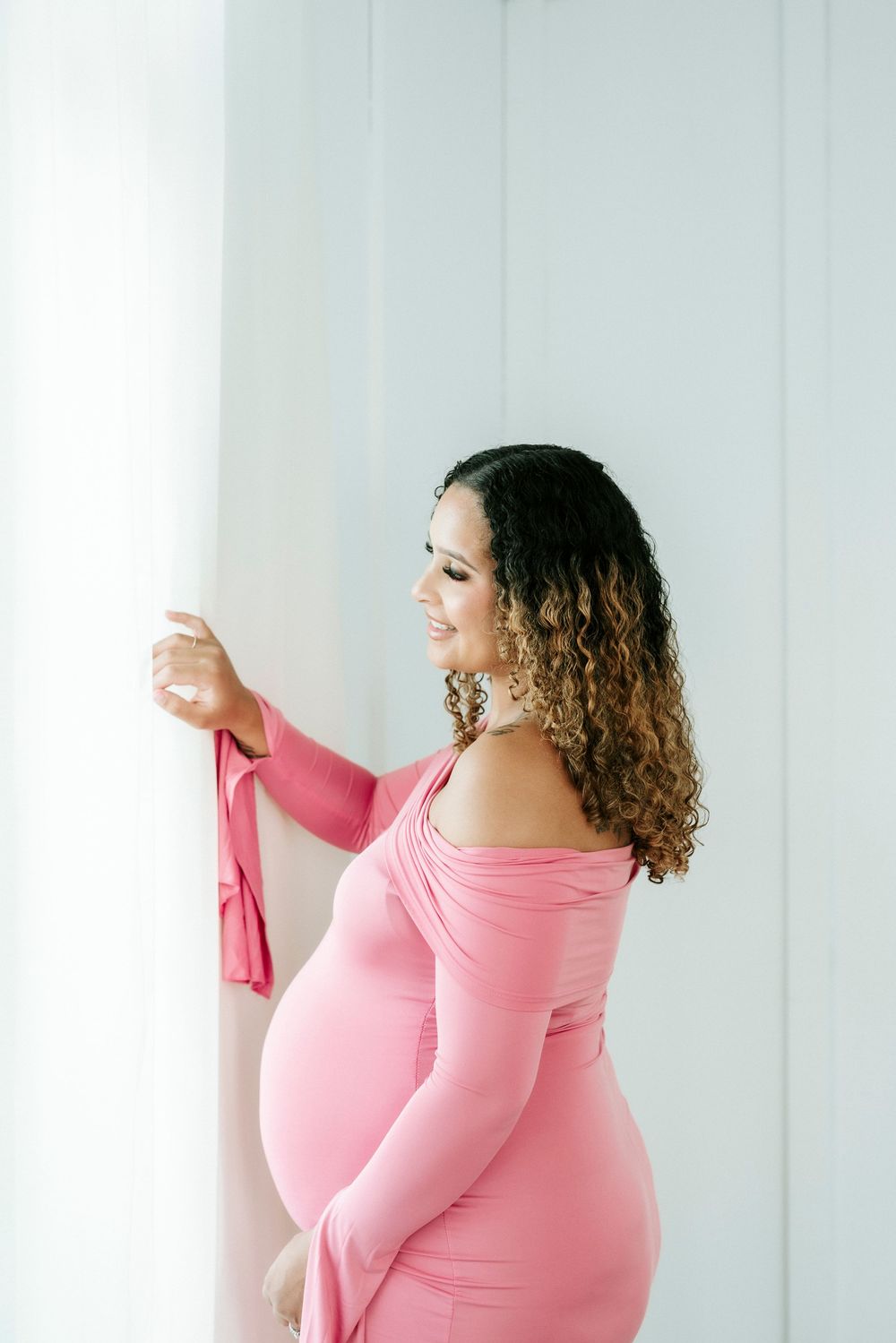 Smiling pregnant woman in pink off-shoulder dress with curly hair, posing by white wall in maternity photo shoot.