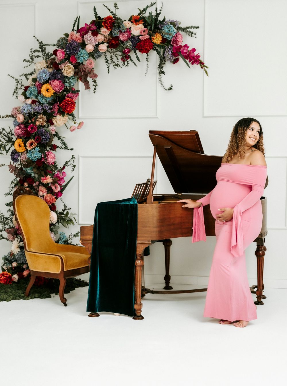 Pregnant woman in pink off-shoulder gown poses by a grand piano with colorful floral arch backdrop.