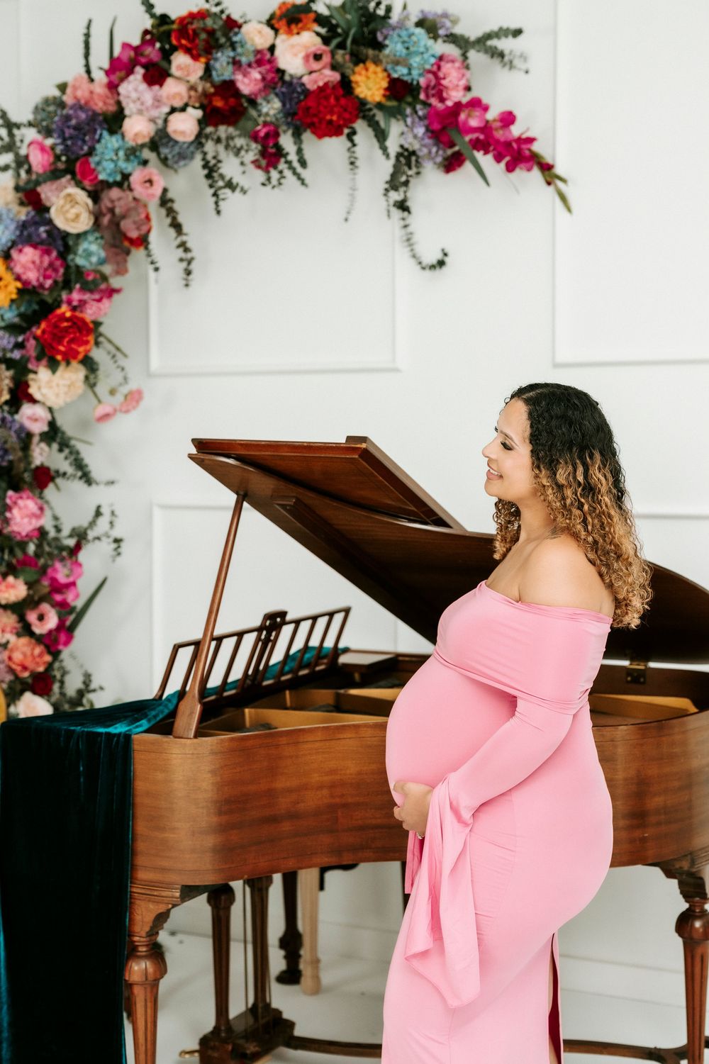 Pregnant woman in pink off-shoulder gown stands by a grand piano, surrounded by colorful floral arch decor.