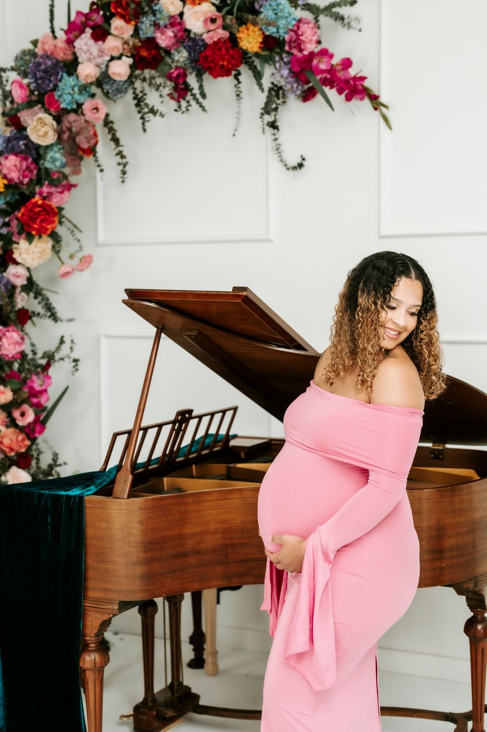 Pregnant woman in pink off-shoulder gown poses by grand piano with colorful floral arch backdrop.