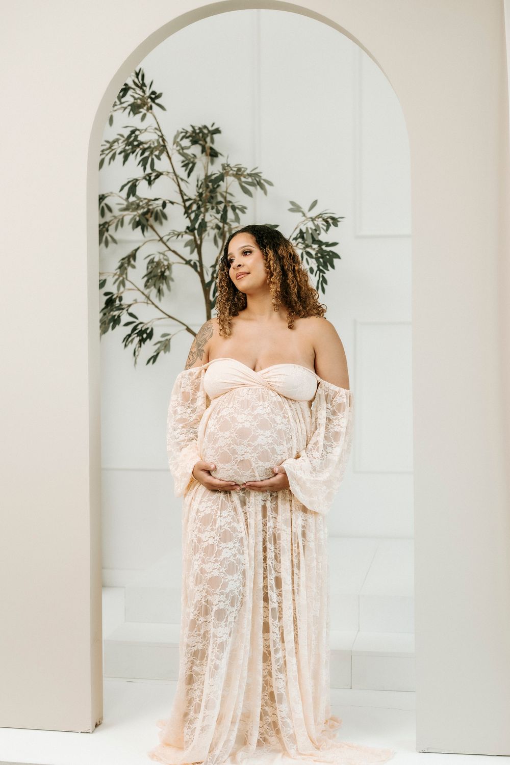 Pregnant woman in blush lace maternity gown cradling baby bump, standing in white arched doorway with greenery.