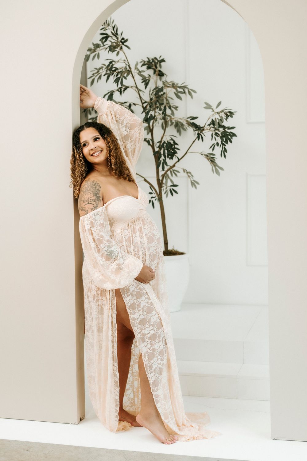 Pregnant woman in blush lace maternity gown poses in white arched doorway with small tree.
