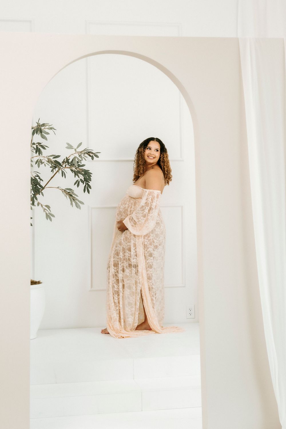 Pregnant woman in elegant blush lace off-shoulder maternity gown standing in white arched doorway with plant.
