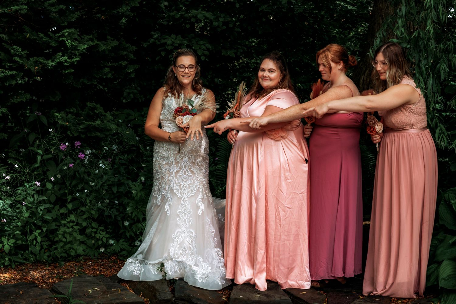 Bride in lace gown with three bridesmaids in pink dresses celebrating outdoors among lush green trees.
