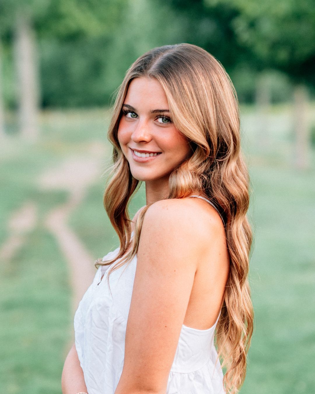 Young woman with long wavy blonde hair smiling outdoors in a white dress with a green bokeh background.