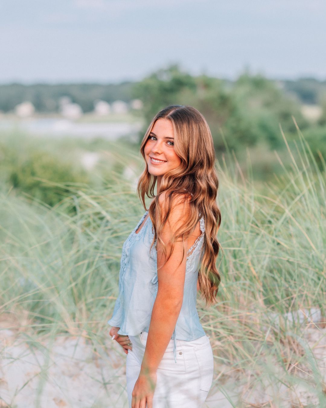 Young woman with long wavy hair smiles over shoulder on sandy beach dunes wearing light blue top and white skirt.