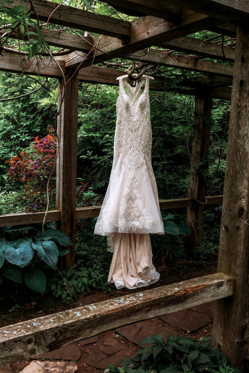 Elegant beaded lace wedding dress hanging in a rustic wooden garden arbor surrounded by lush green foliage.
