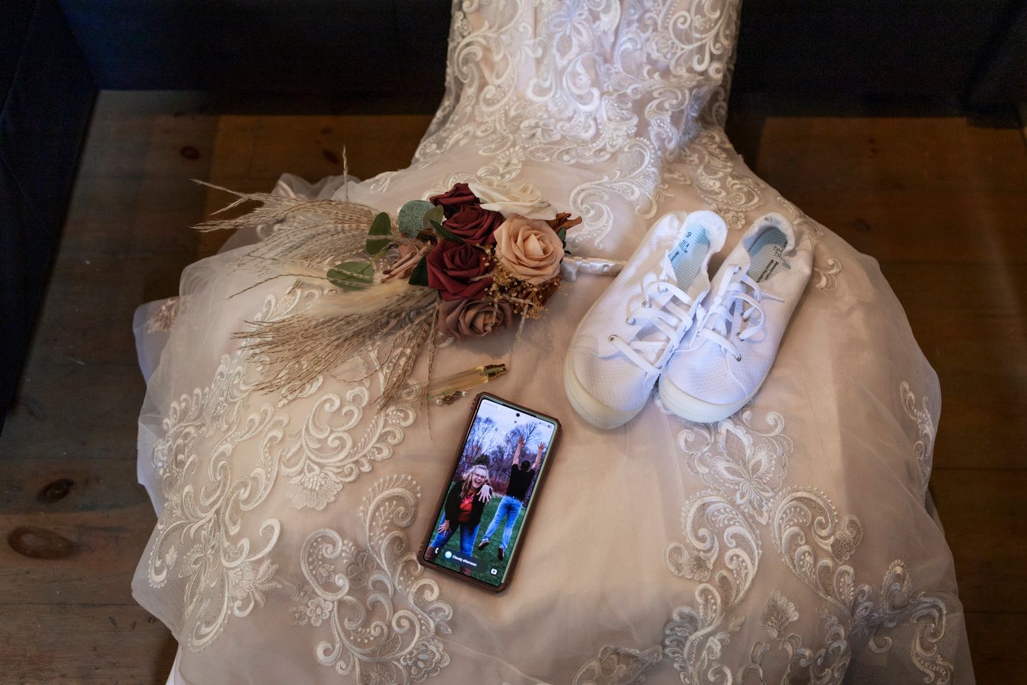 Wedding dress laid flat with white sneakers, floral bouquet, and phone displaying couple photo on wooden surface.