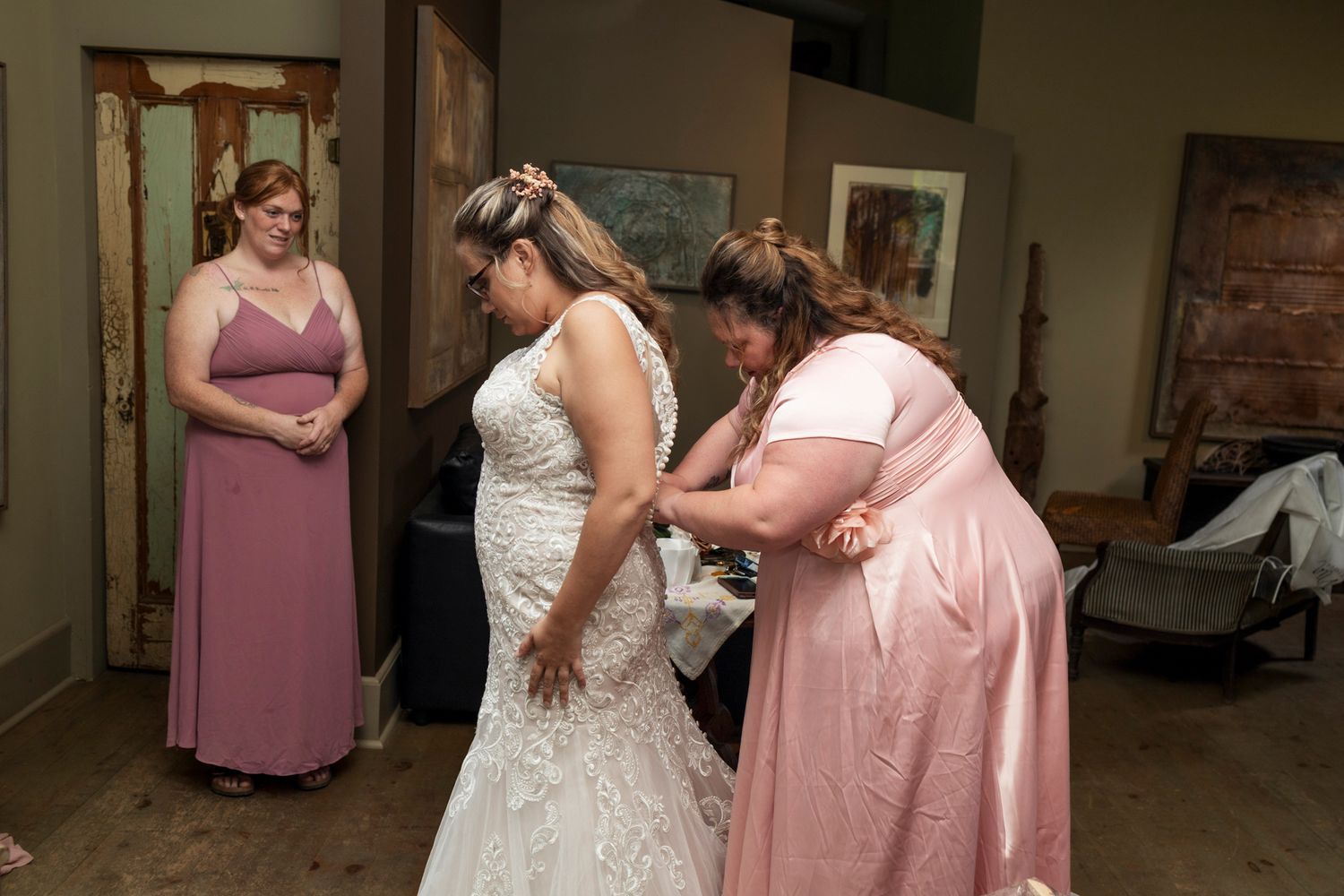 Bridesmaids in pink dresses help a bride in a white lace gown get ready in a rustic indoor venue.