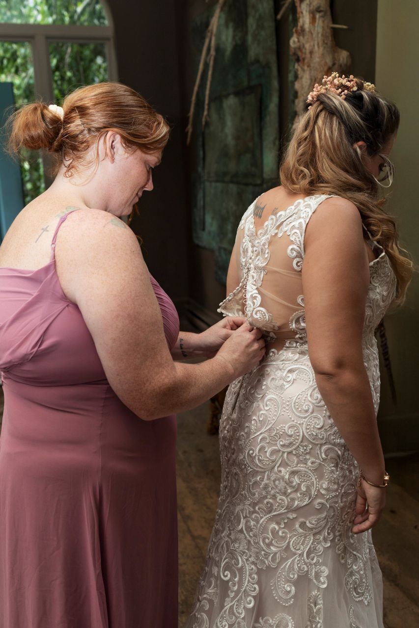 Bridesmaid in pink dress fastening the back of a brides lace wedding gown before the ceremony.