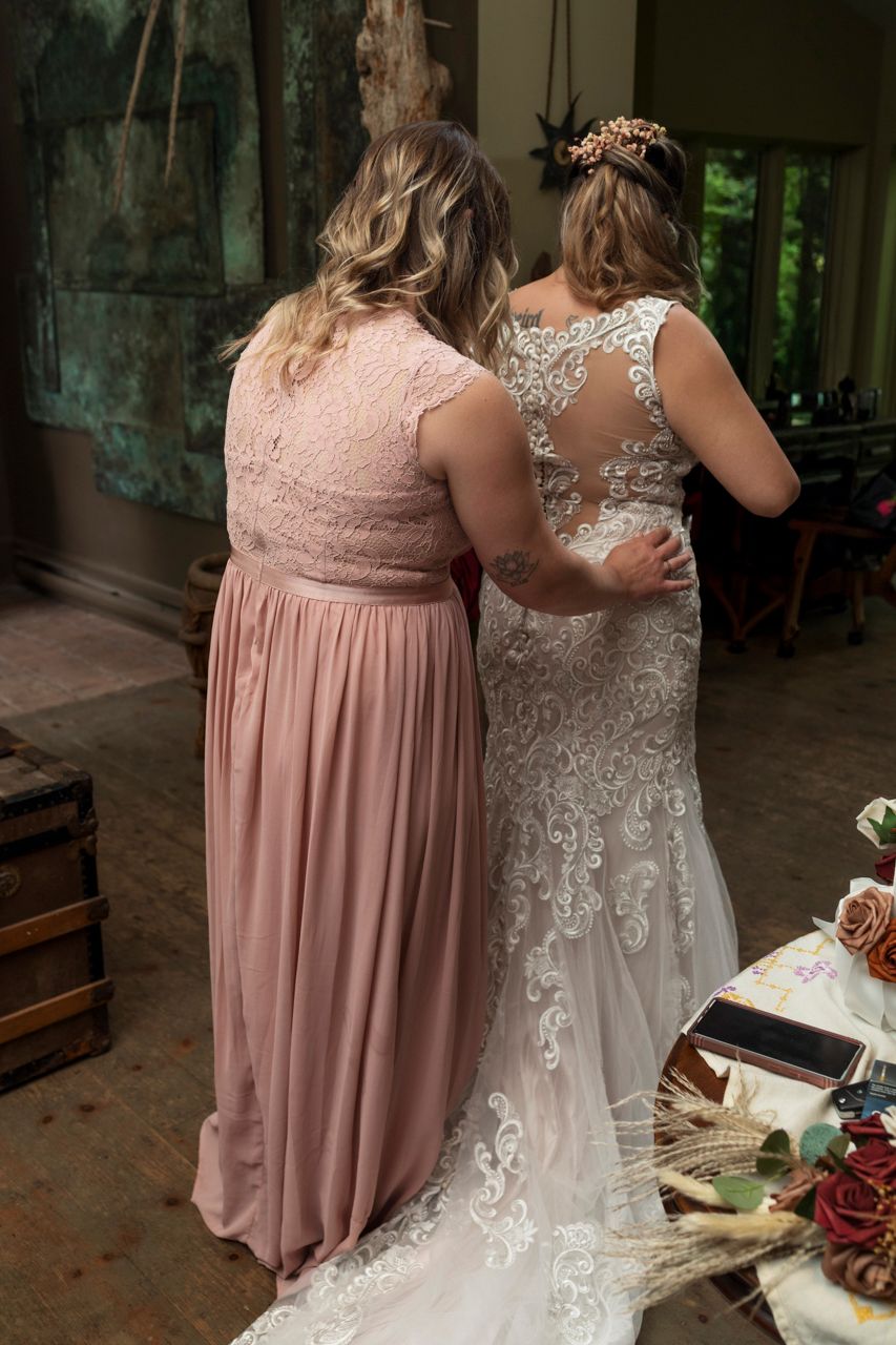 Bride in lace wedding gown with bridesmaid in dusty rose dress standing together at wedding venue.