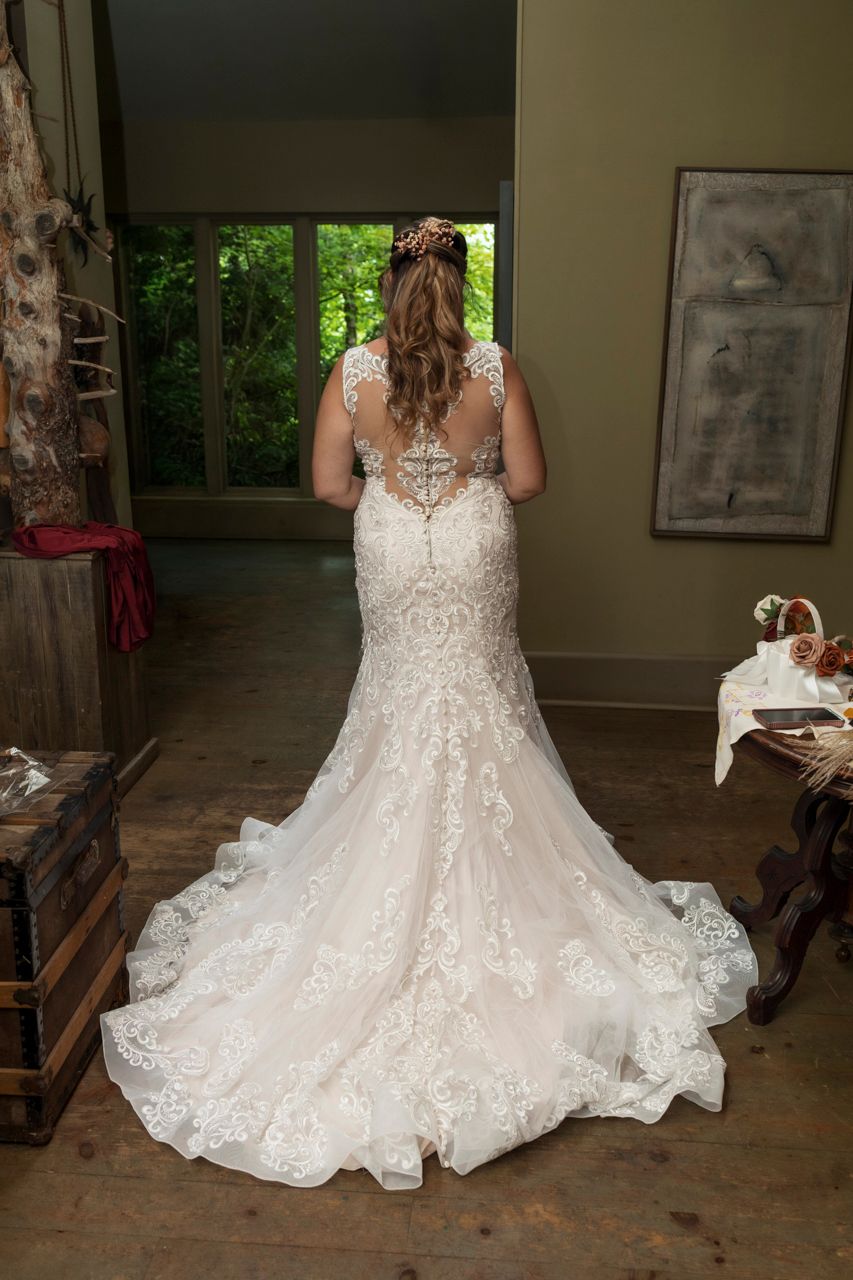 Bride in elegant lace mermaid wedding gown with illusion back and long train, viewed from behind near a window.