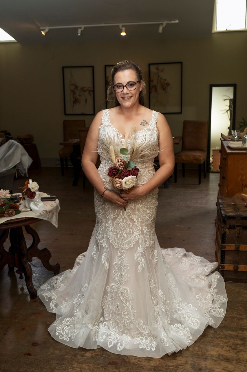 Bride in lace mermaid wedding gown holding burgundy bouquet stands in elegant venue.