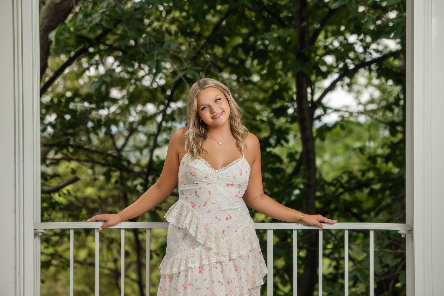 Young woman in floral dress posing on white balcony surrounded by lush green trees.