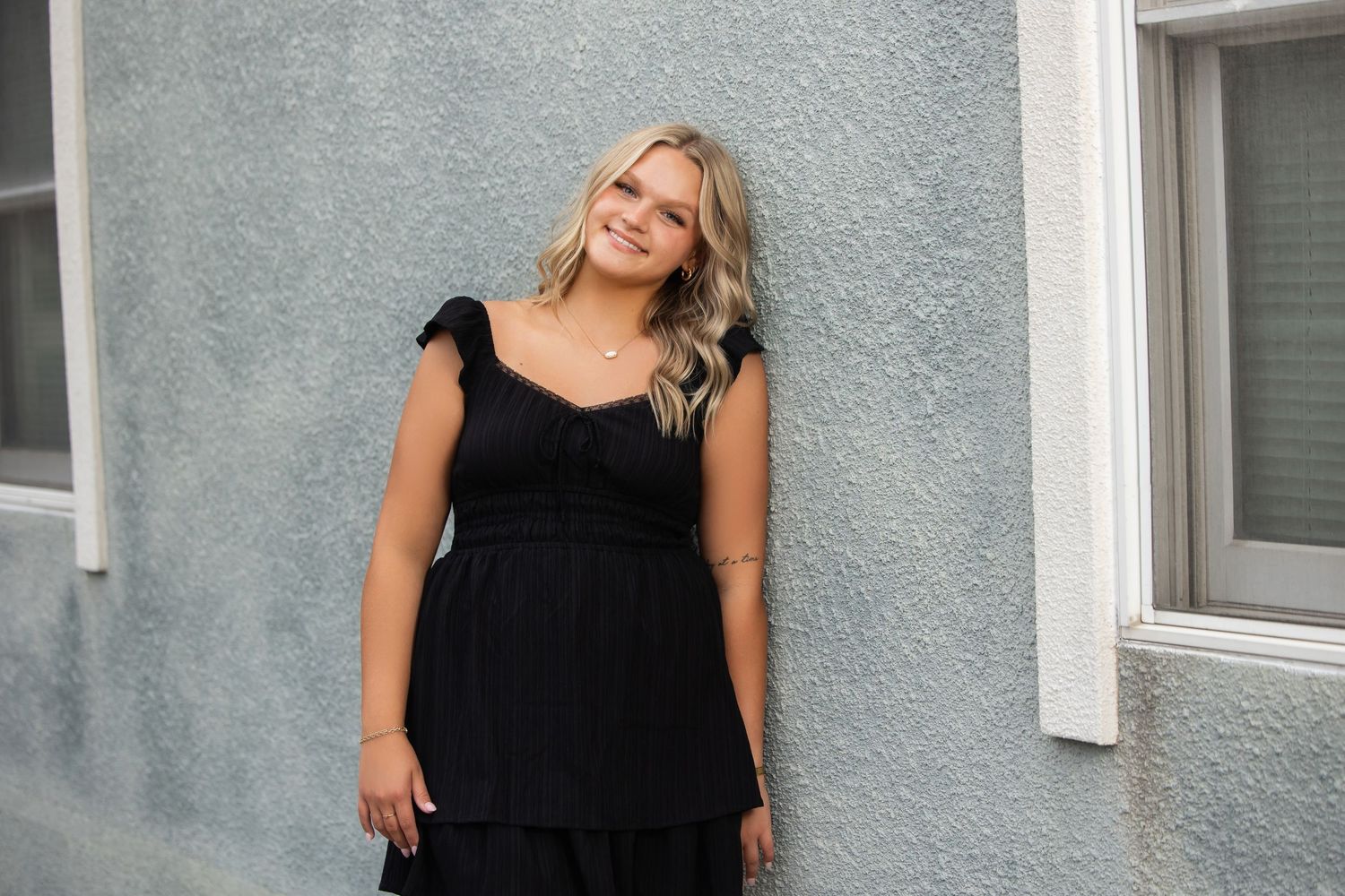 Young woman in black dress smiling against gray wall outdoors.