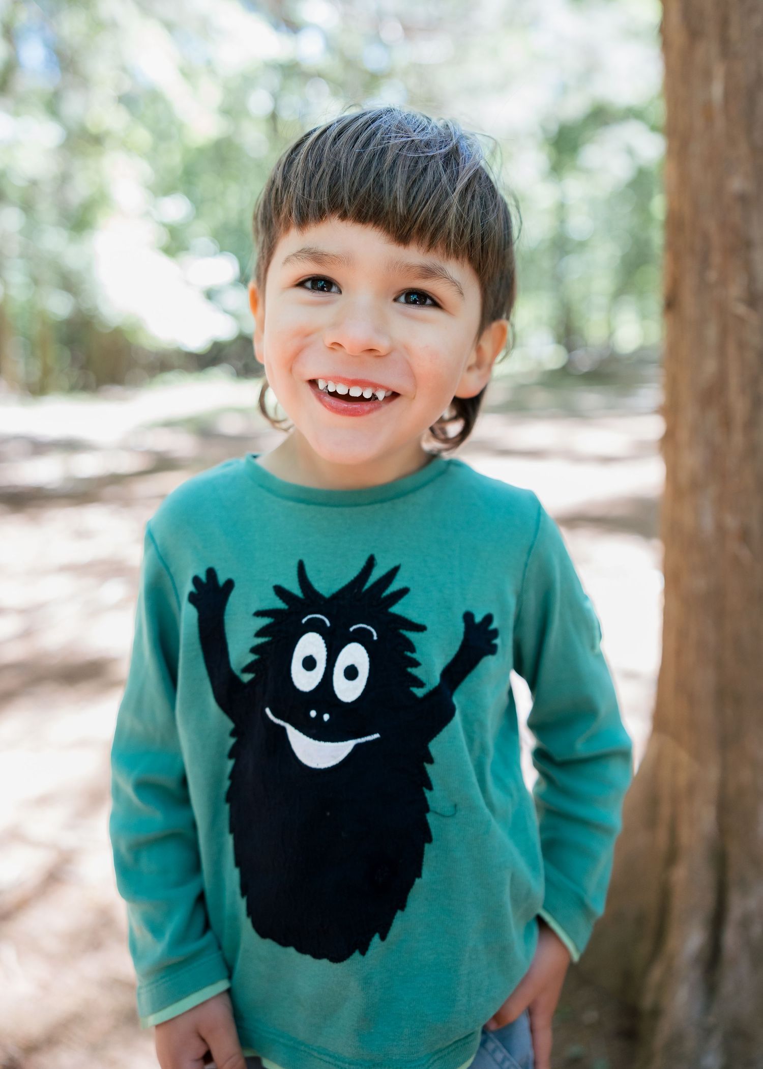 Smiling young boy wearing a teal long-sleeve monster graphic shirt, standing outdoors among trees.