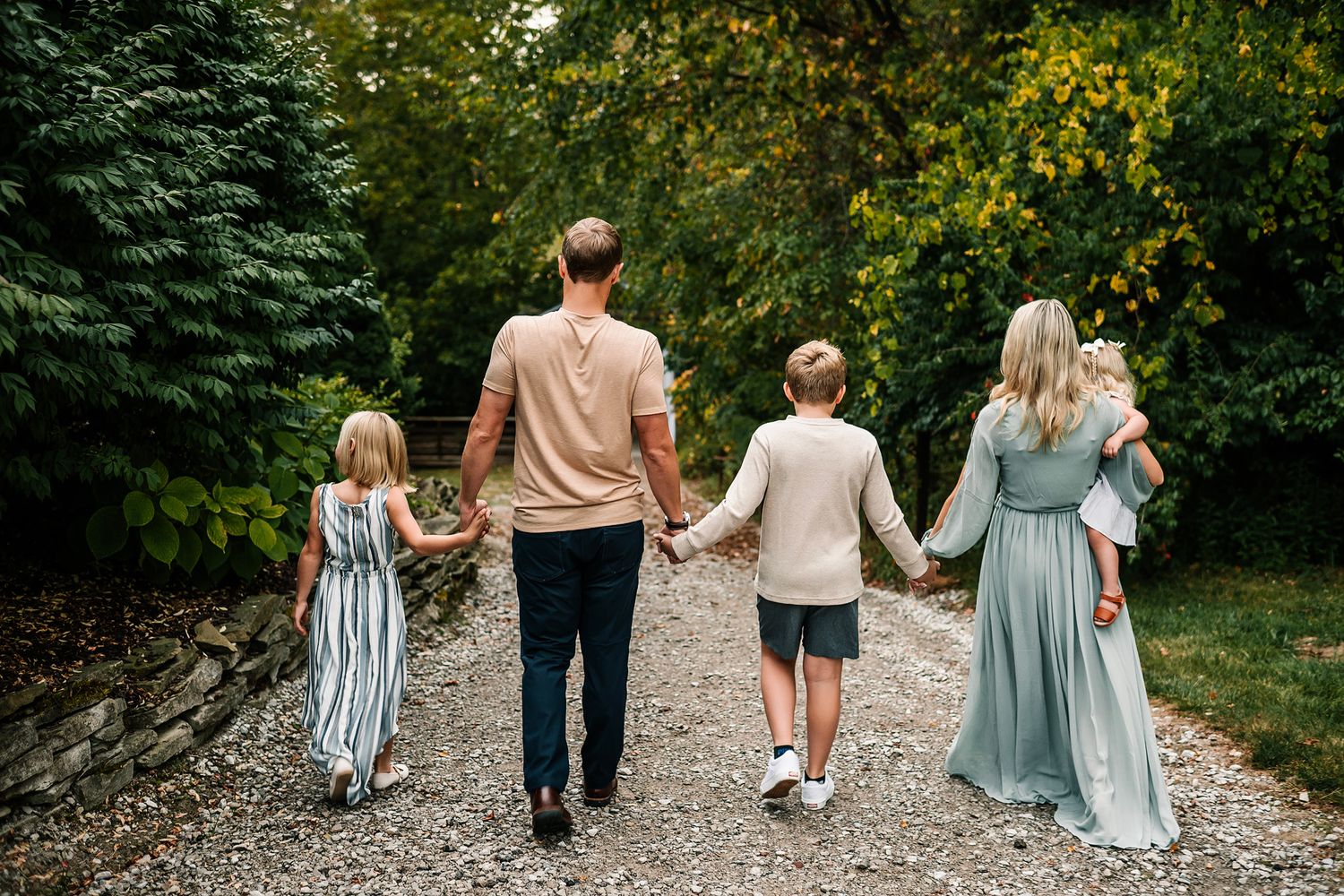 Family of five walking hand-in-hand down a gravel path surrounded by lush green trees.