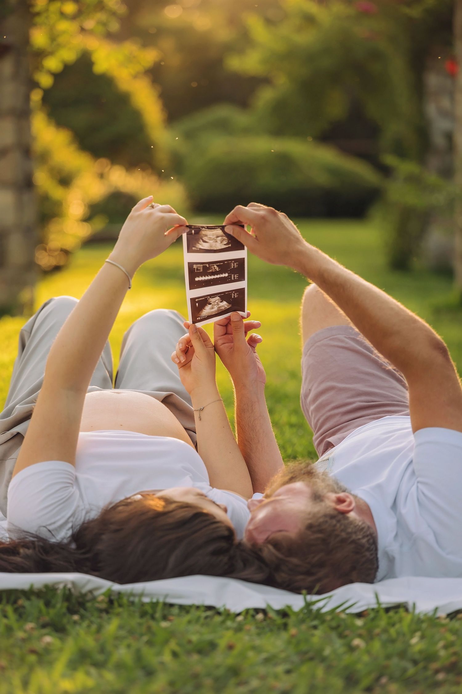 Pregnant couple lying on grass in park, holding ultrasound photos up together in golden sunlight.