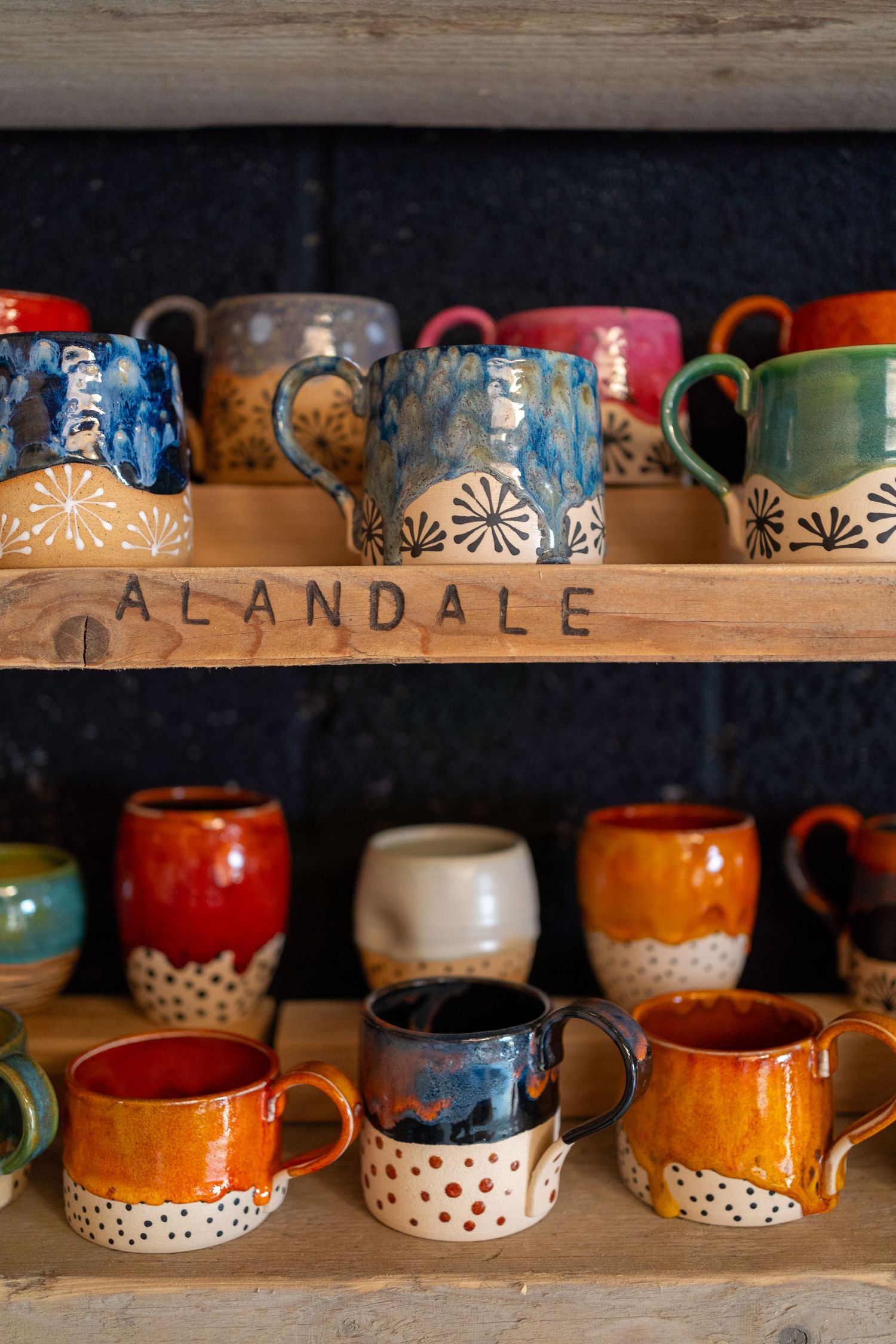 A woman with gray hair and glasses holds a colorful mug, wearing a green shirt and pink skirt near a shelf of ceramics.