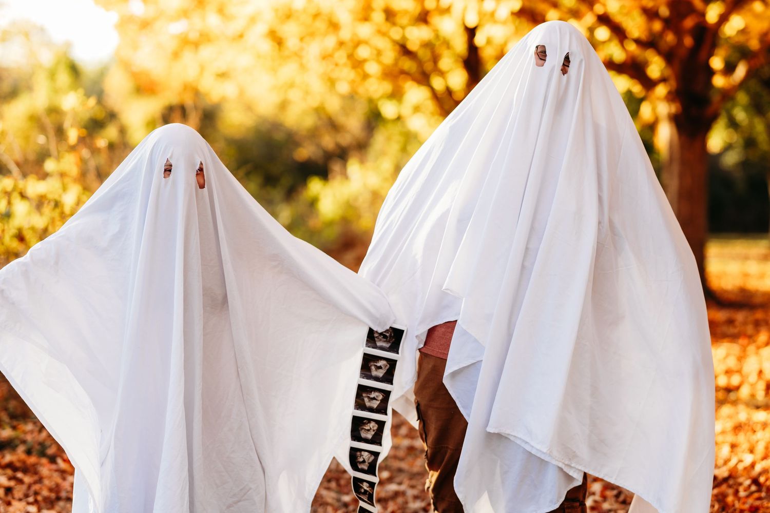 Two children in white ghost costumes trick-or-treating outdoors among autumn trees on Halloween.