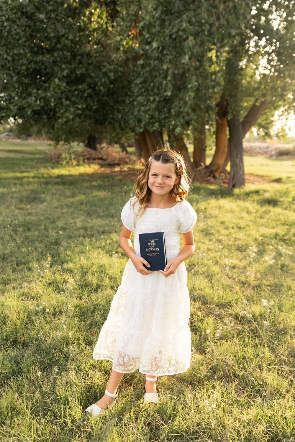 Young girl in white dress holding a Book of Mormon, standing on green grass under trees in golden sunlight.