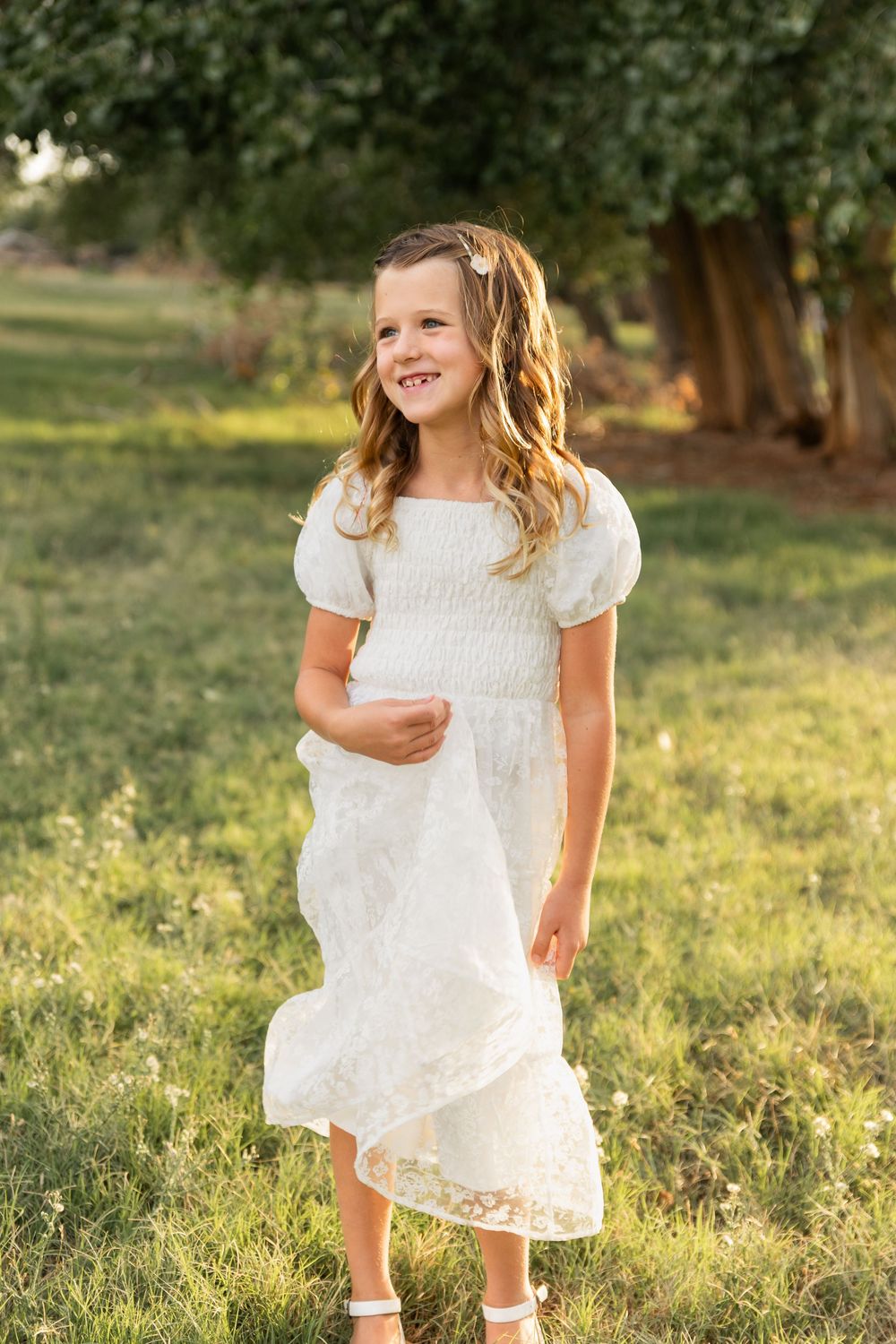 Young girl in white lace dress smiling outdoors in green park setting during golden hour.