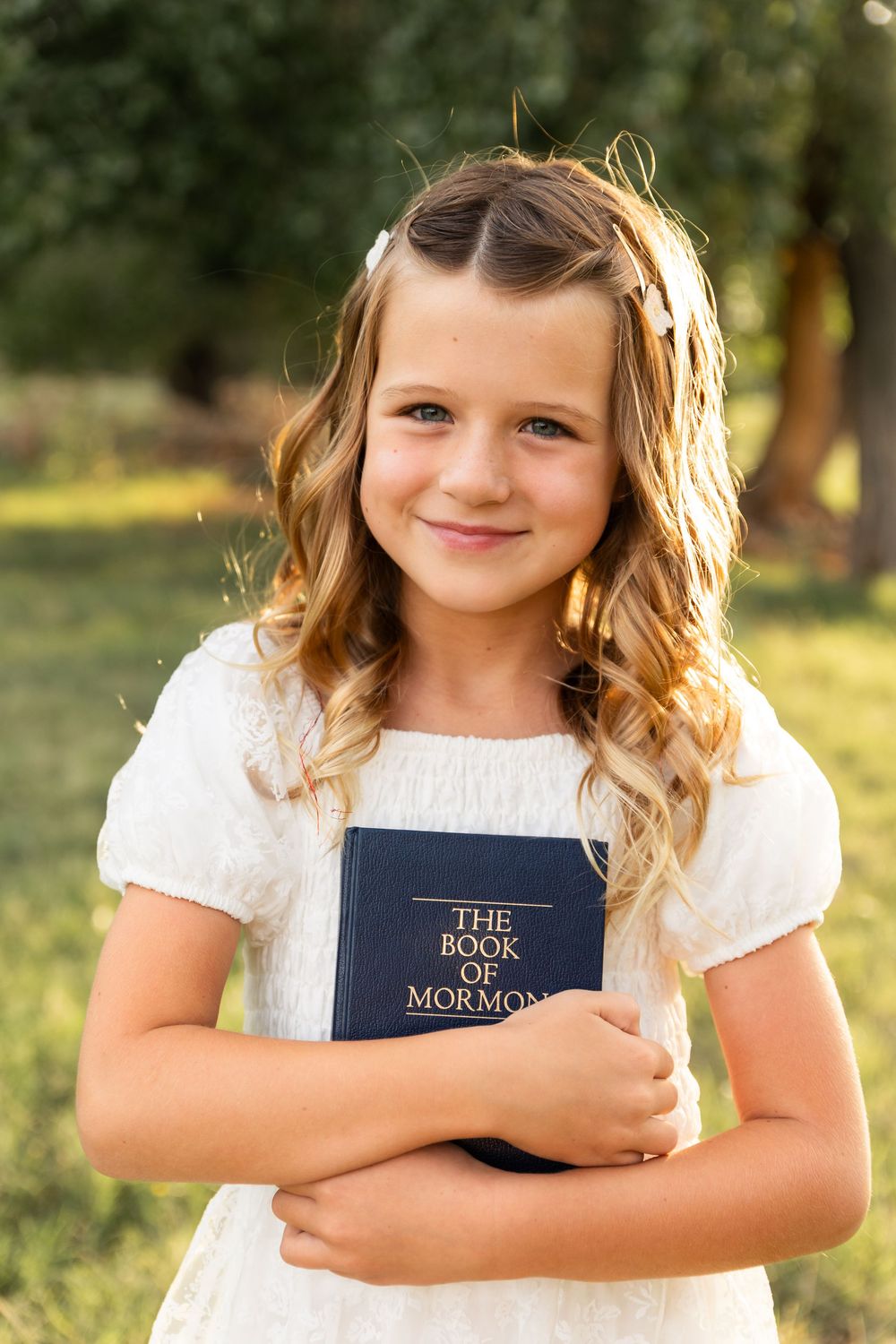 Young girl in white dress holding Book of Mormon scripture outdoors in natural light.