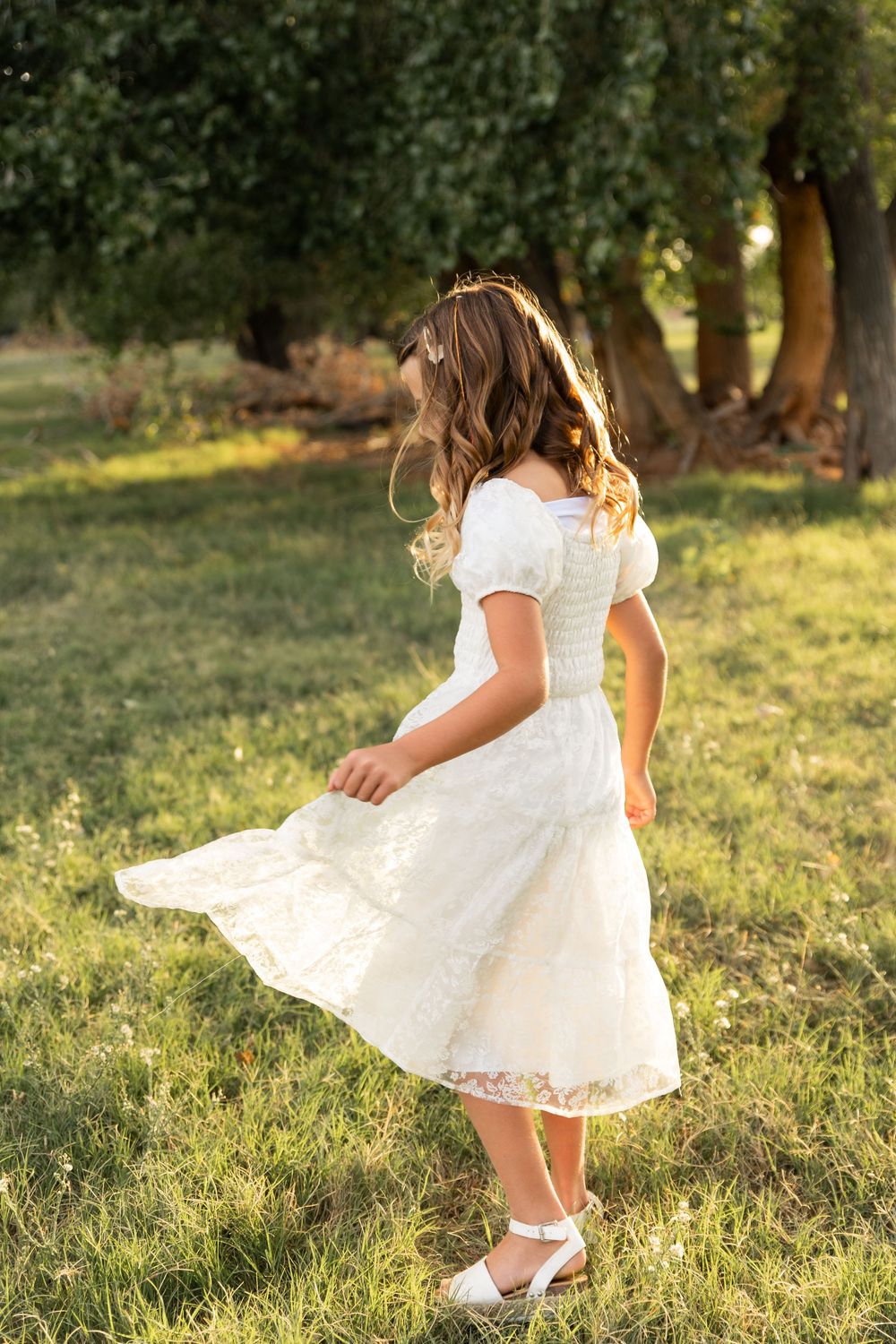 A young girl in a white lace dress twirls gracefully in a sunlit park surrounded by green trees.