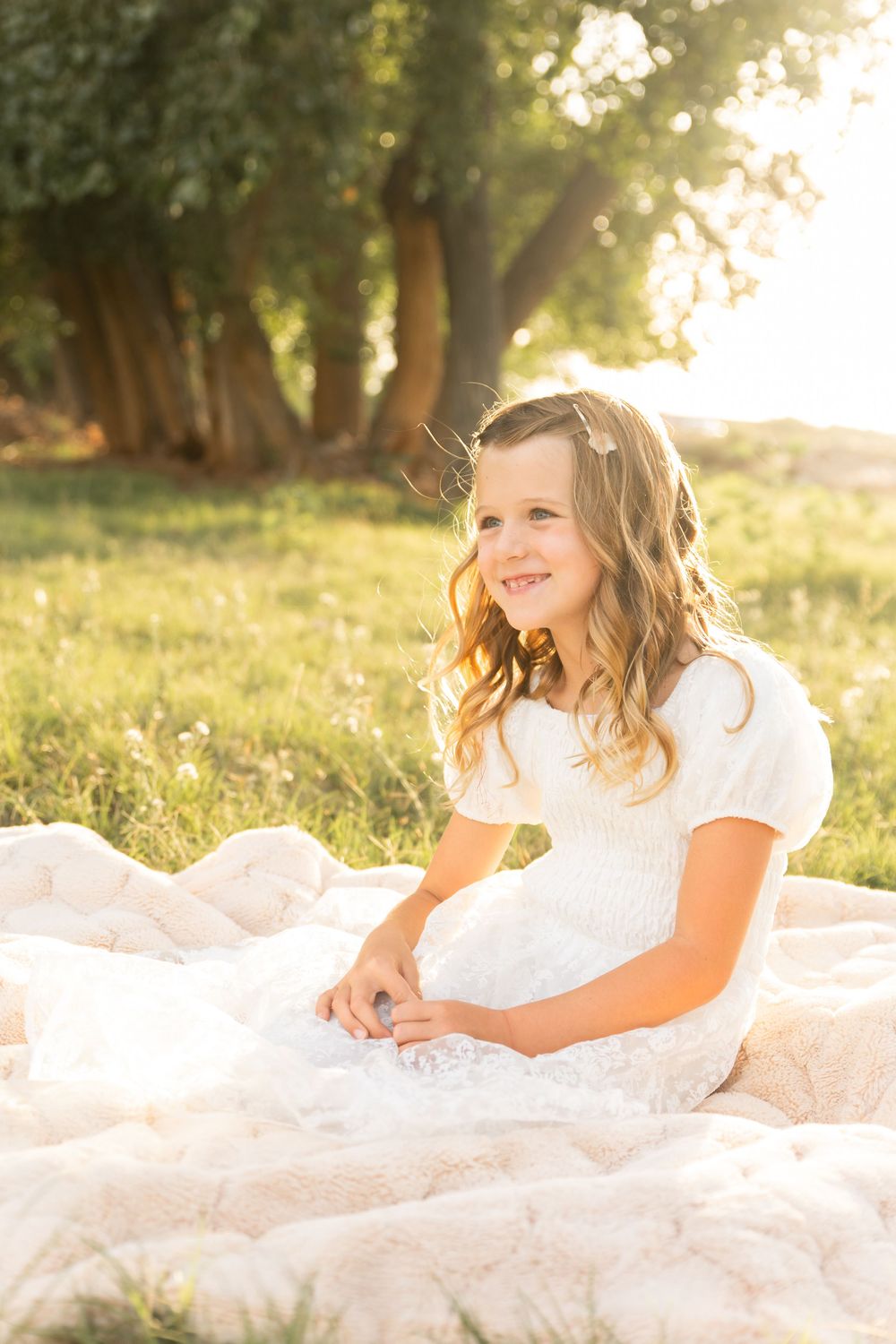 Young girl in white lace dress sitting on blanket in sunlit meadow, smiling joyfully with trees in background.