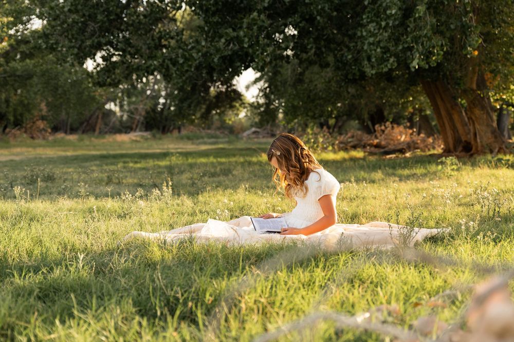 Young woman sitting on blanket in sunny green field, reading a book under a large tree in golden afternoon light.