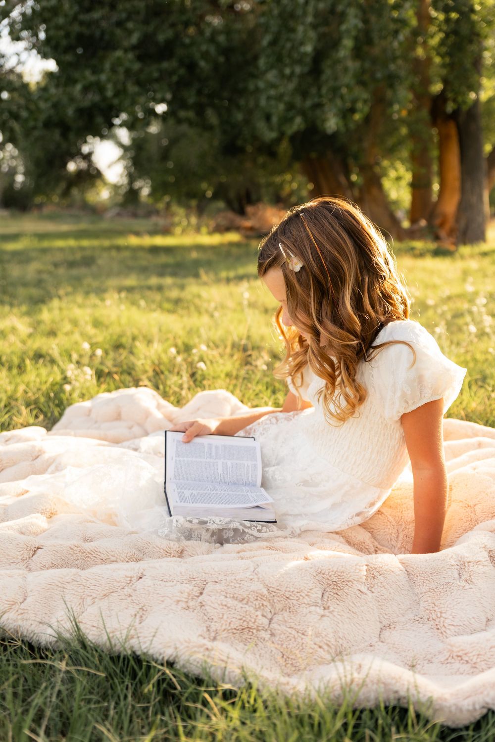 Young girl in white dress reading a book on a blanket in a sunny green park.