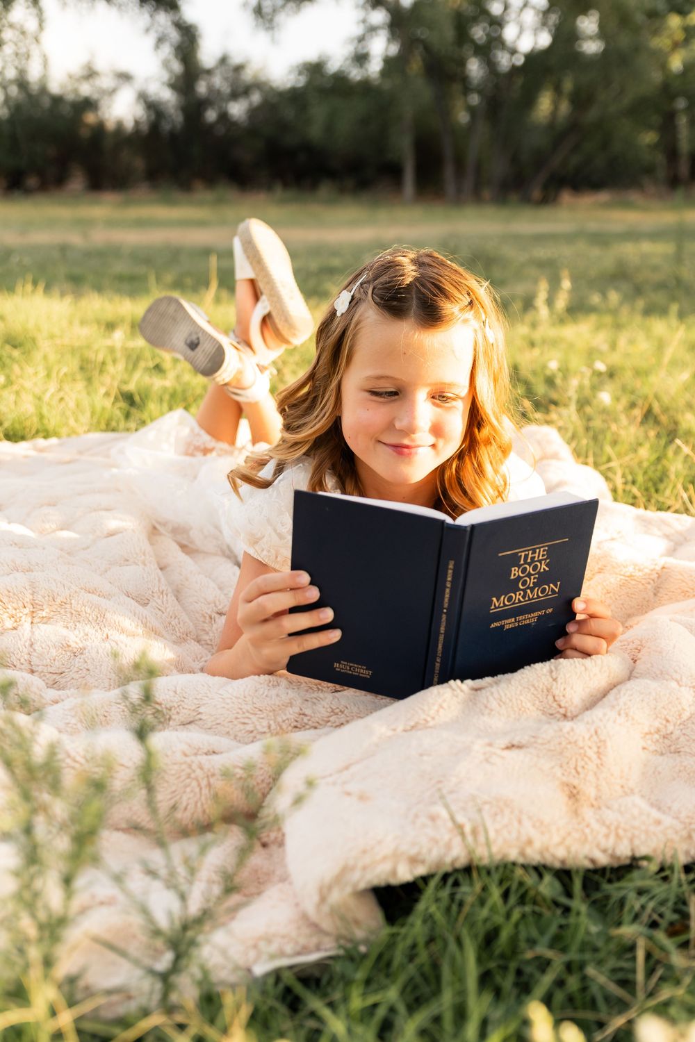 Young girl lying on a blanket outdoors reading the Book of Mormon in a sunny green field.
