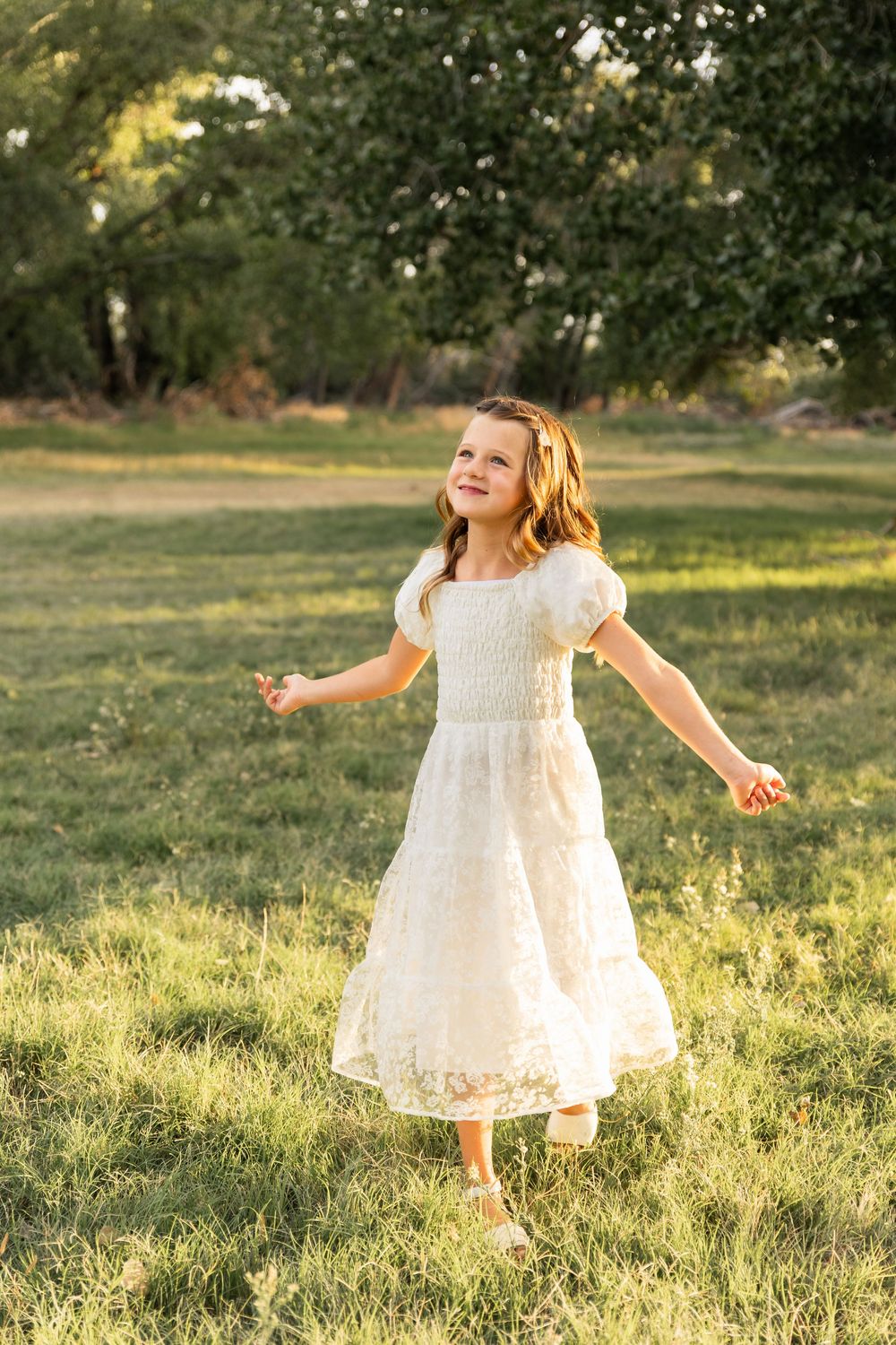 A young girl in a white lace dress twirls joyfully in a sunlit green field with trees in the background.