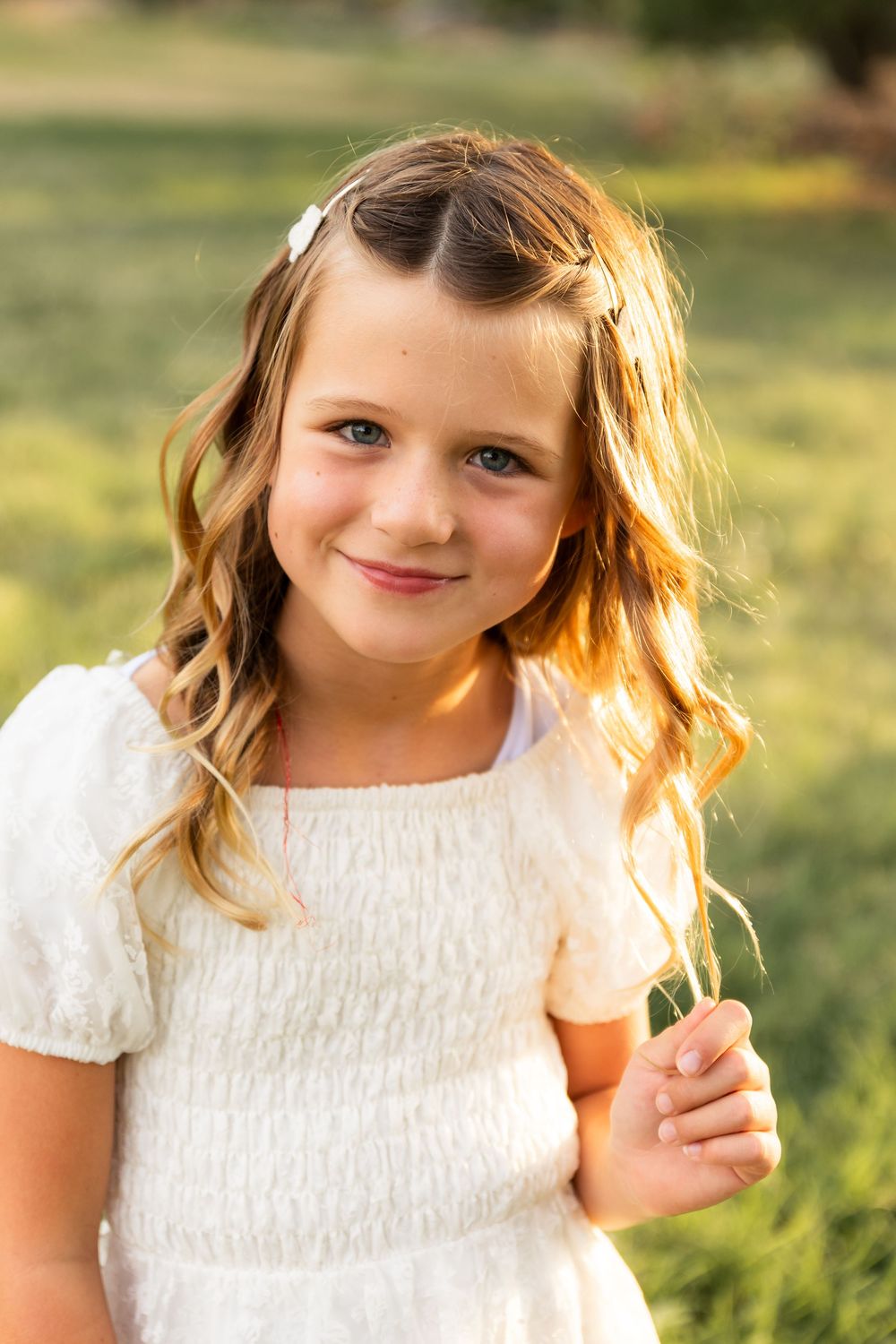 Young girl in white dress with curly hair smiling outdoors in golden sunlight.