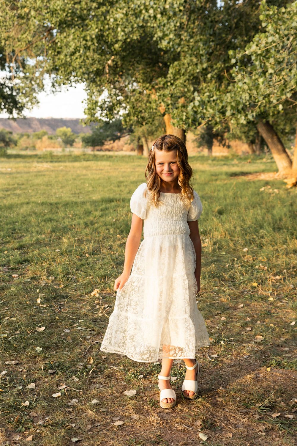 Young girl in white lace dress and sandals standing in sunlit park with green trees.