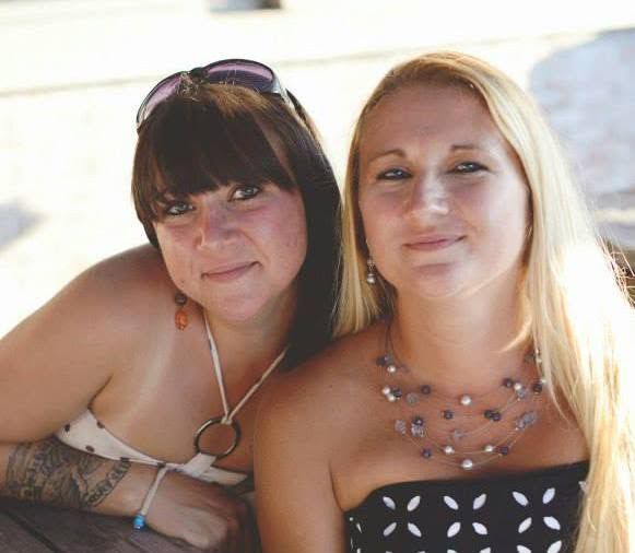 Two women posing together outdoors, one with dark hair and sunglasses, the other blonde wearing a floral strapless top.