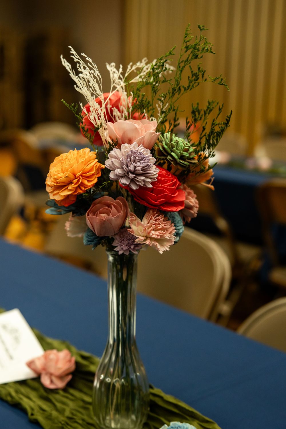 Colorful floral centerpiece in tall glass vase with orange, red, purple blooms on blue tablecloth at event.