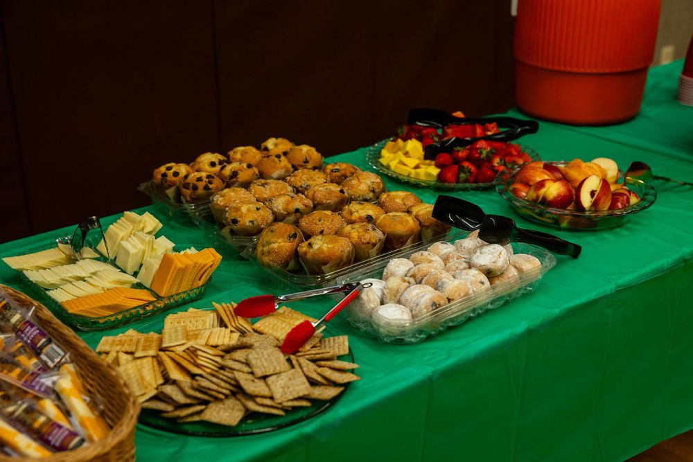 A buffet table with green cloth displays muffins, cheese, crackers, fruit, and round desserts at a catered event.