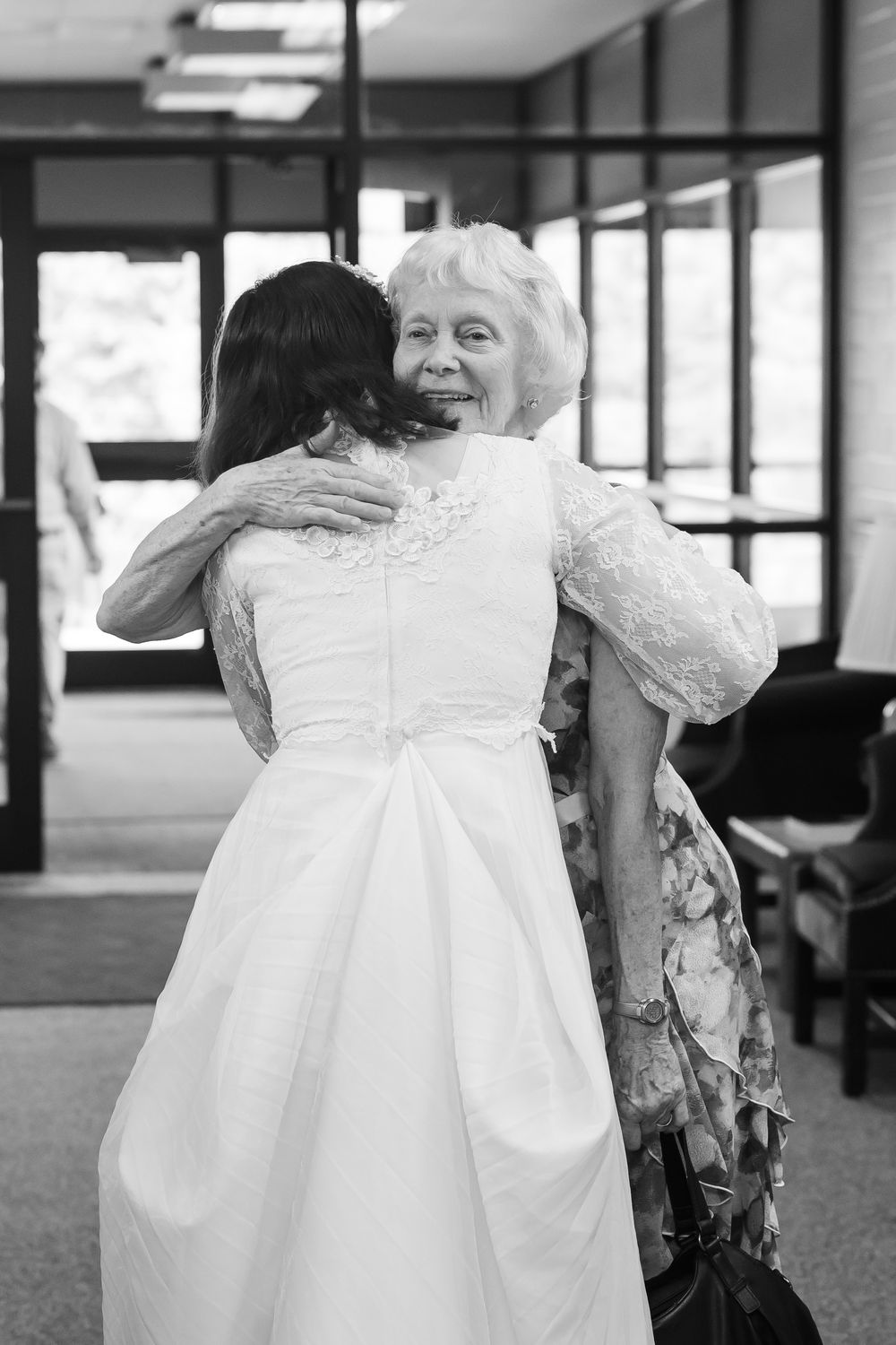 Bride in white gown hugging elderly woman in lace dress at wedding venue, black and white photo.