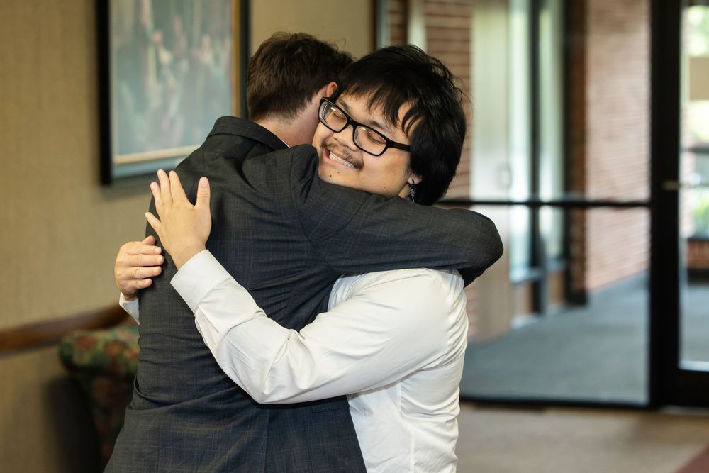 Two men sharing a warm hug, one smiling broadly wearing glasses and a white shirt indoors.