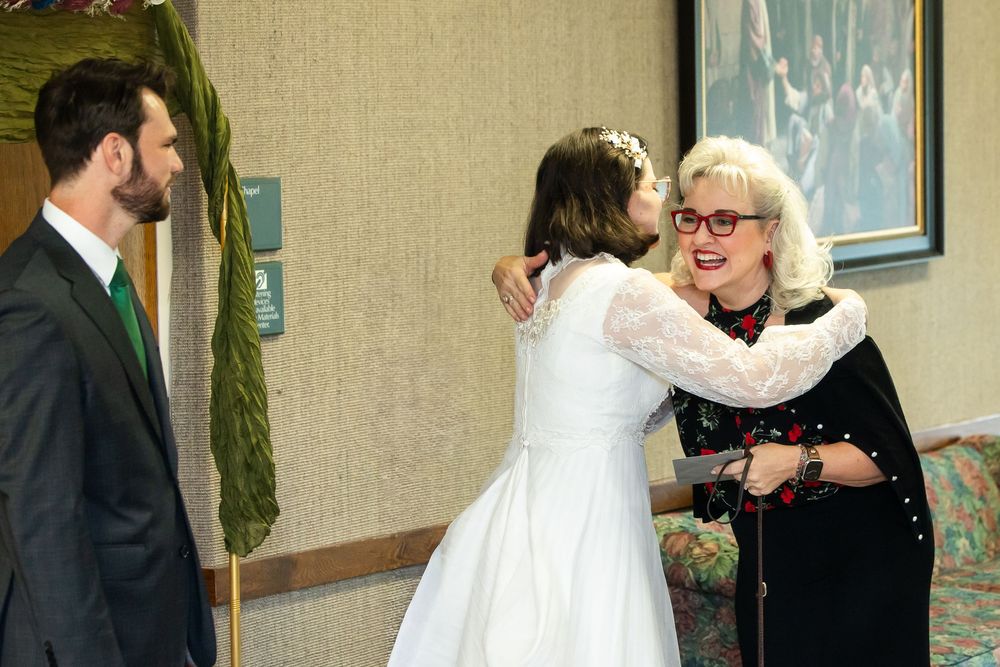 A bride in white dress hugs a woman in red glasses at a wedding reception while a groom in suit watches nearby.