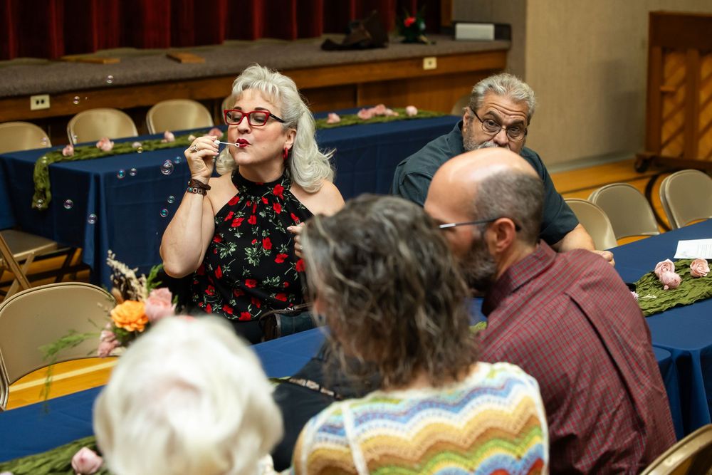 Woman with gray hair and red glasses speaks at an indoor event with attendees seated at blue tables.