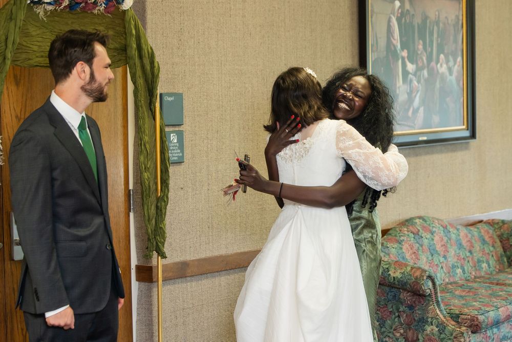 A bride in a white gown joyfully hugs a guest while a man in a suit smiles nearby at a wedding venue.