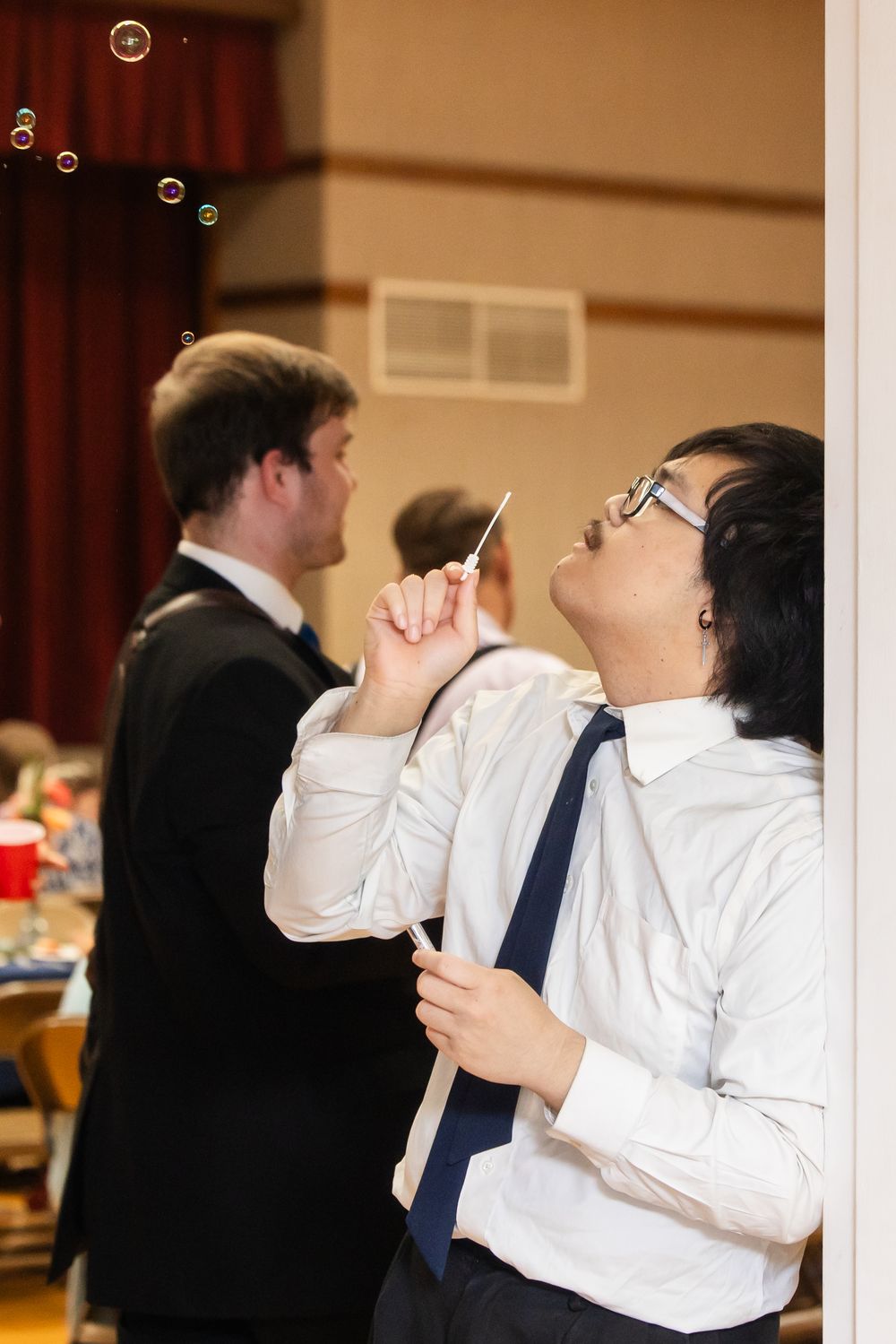 Young man in white shirt and tie blowing bubbles at a formal event, with another man in a suit in the background.