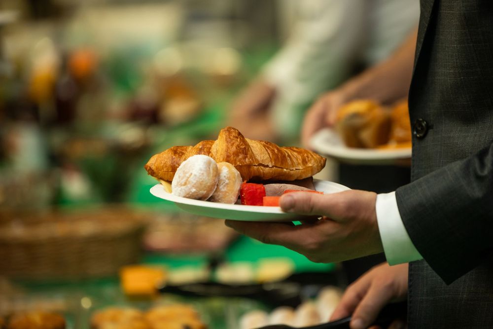 A waiter in a suit holds a white plate with croissants and pastries at an elegant buffet spread.