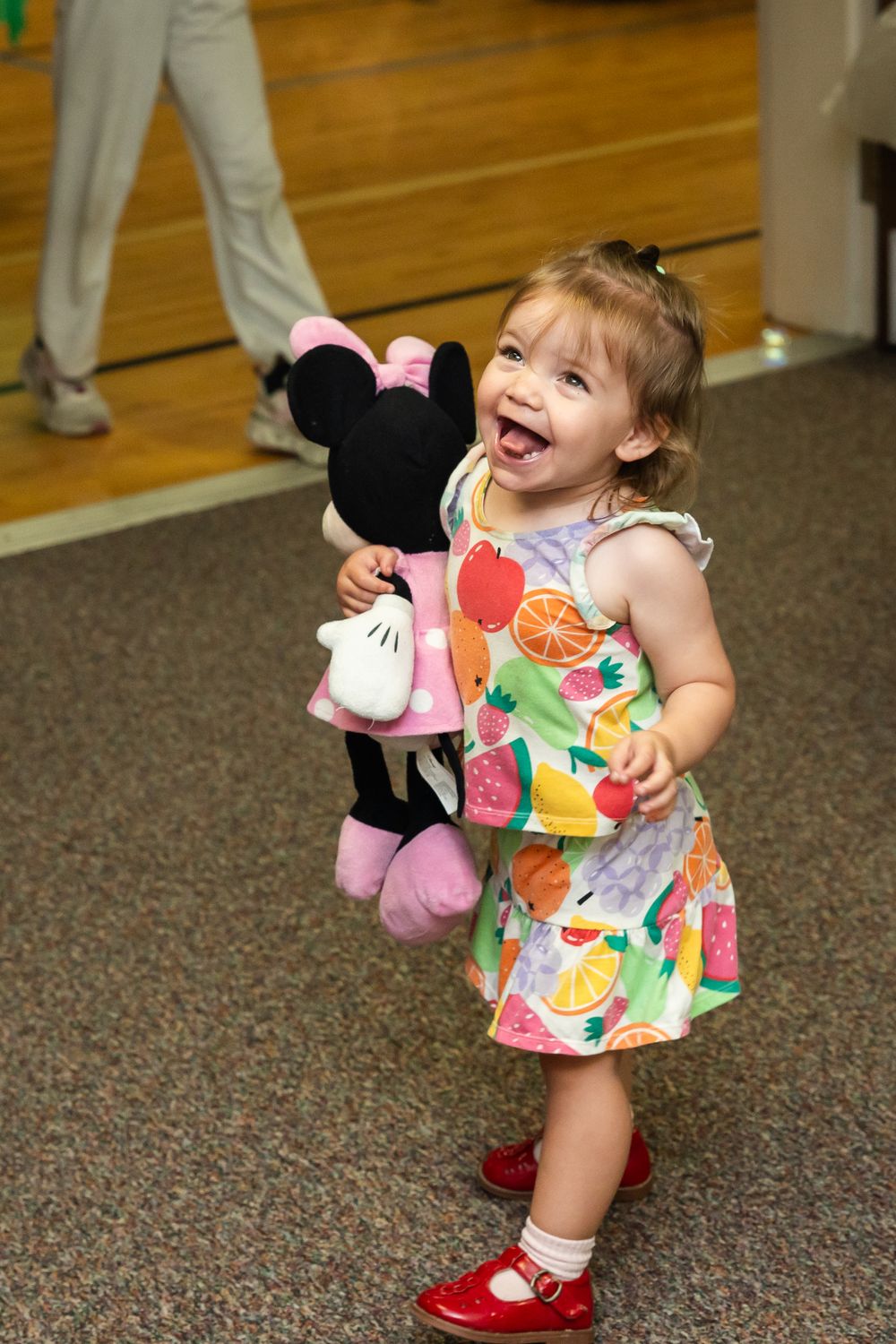 Laughing toddler girl in colorful fruit dress and red shoes holding a Minnie Mouse plush toy.
