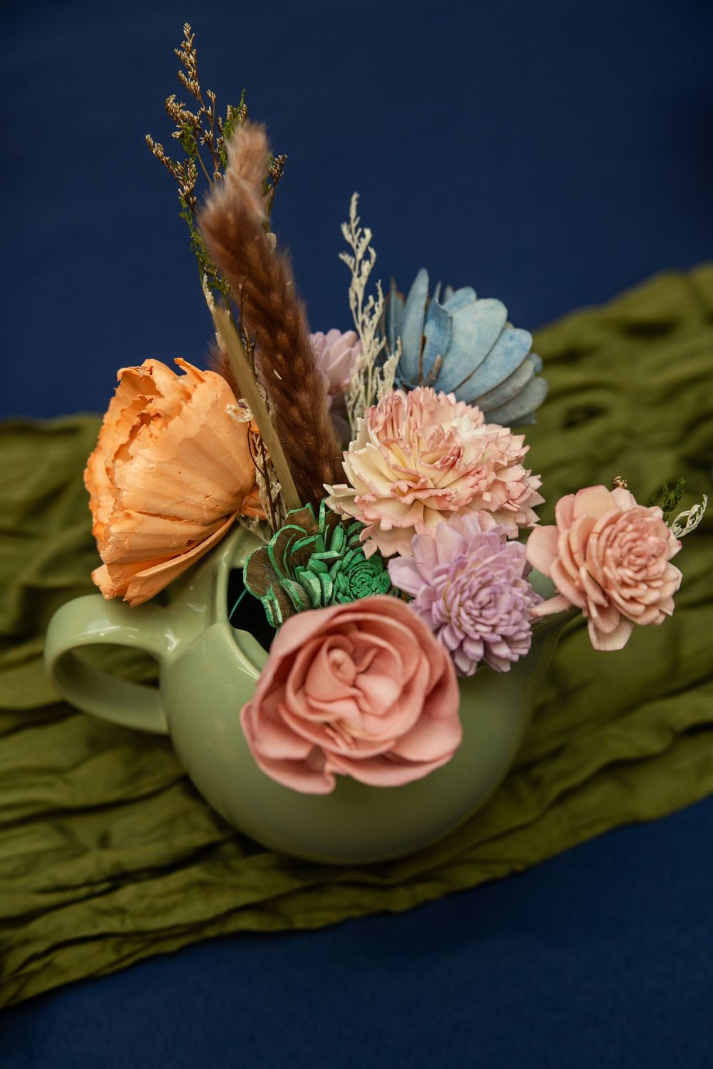 Pastel dried floral arrangement in a sage green teapot vase on olive fabric, featuring roses, carnations, and pampas grass.
