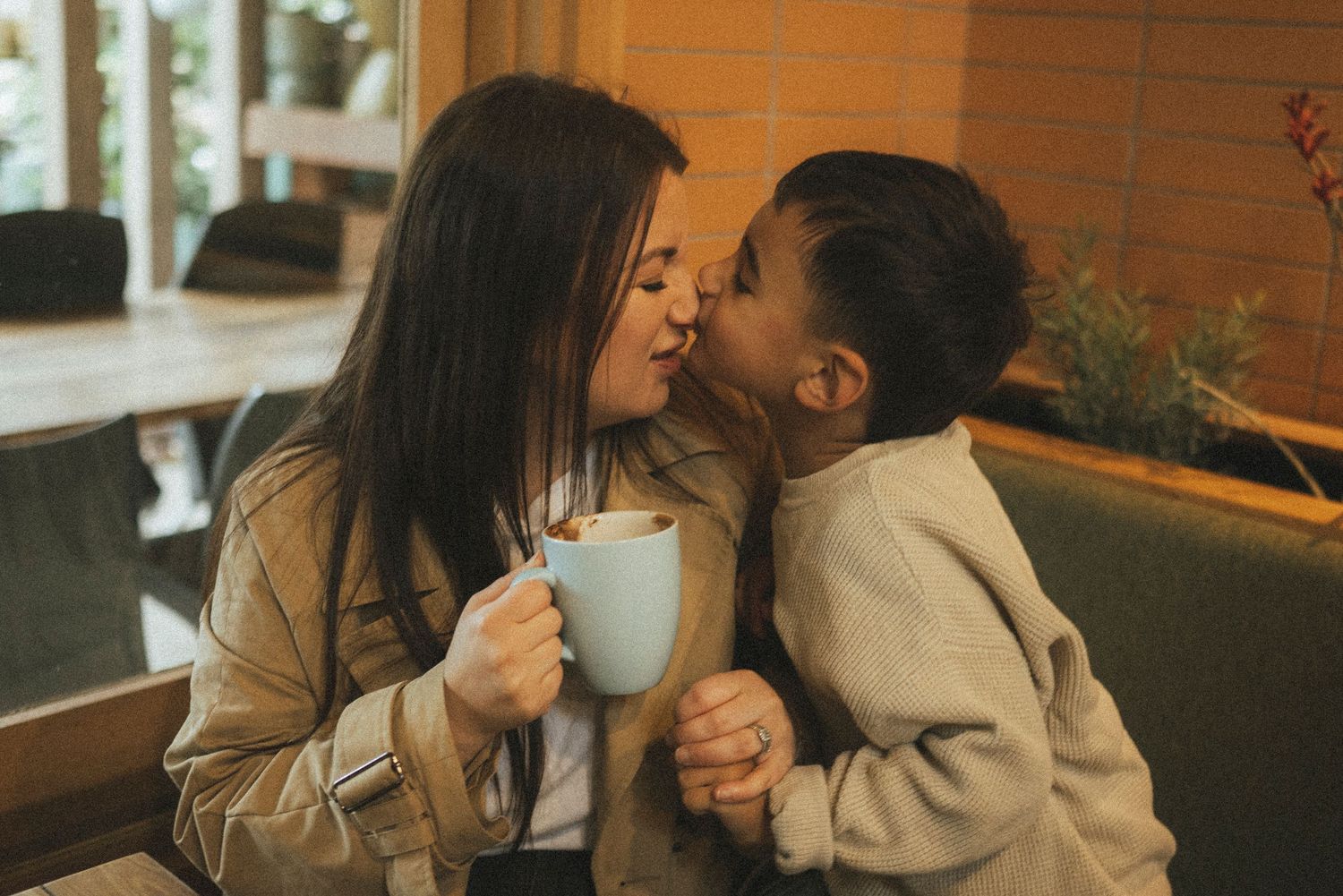 A mother and young child share a tender kiss at a cozy cafe, with the mom holding a warm cup of coffee.