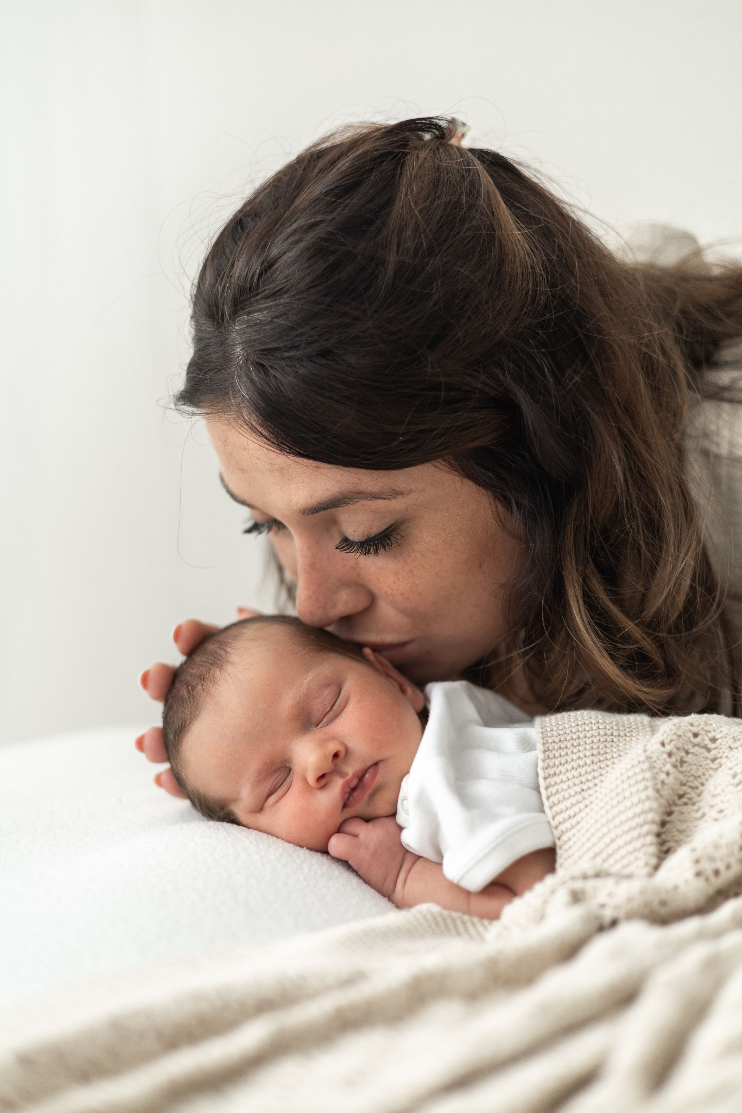 A tender moment as a mother gently kisses her sleeping newborn baby wrapped in a white blanket natural baby photography Nottingham