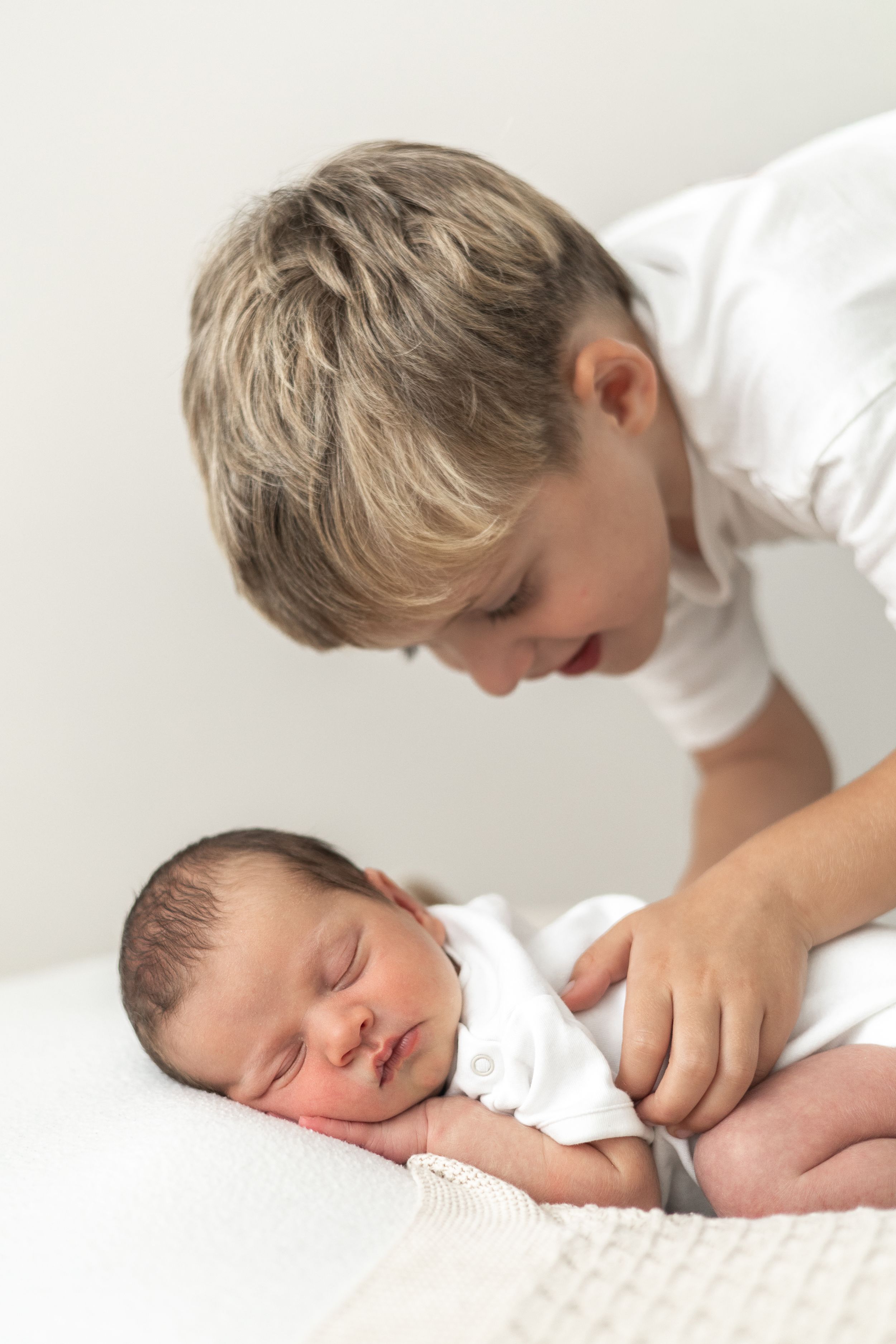 Sibling in white shirt tenderly watching over sleeping newborn baby sibling dressed in white natural baby photography Nottingham